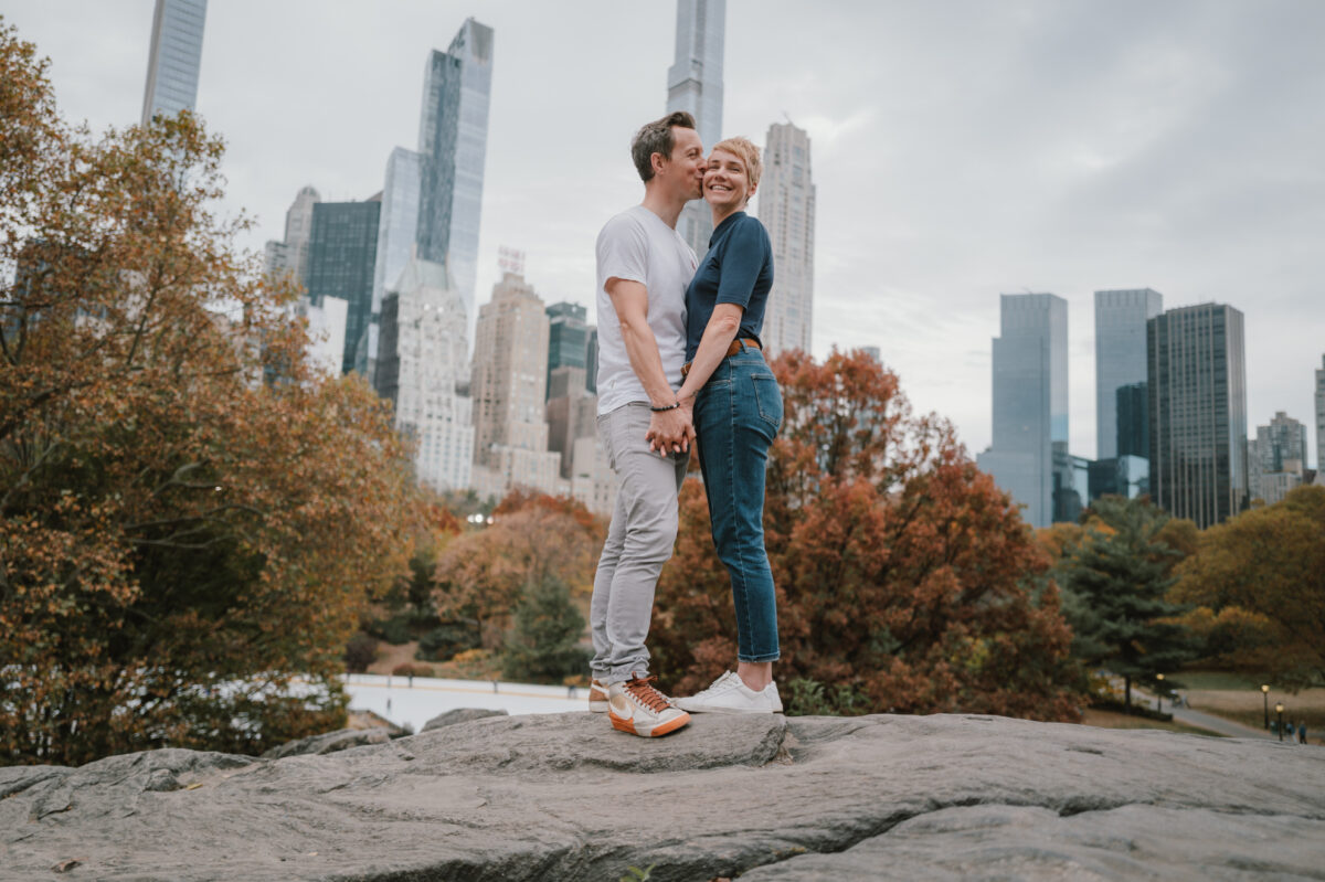    Central Park engagement photos in autumn, kiss on the cheek with the city behind